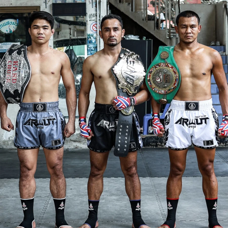 Three Muay Thai fighters standing side by side, each holding championship belts, with a building and street in the background.