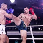 A dynamic action shot of a Muay Thai or boxing match in a ring, featuring two fighters. One fighter in blue shorts and red gloves delivers a high kick, while the other fighter in white shorts defends.