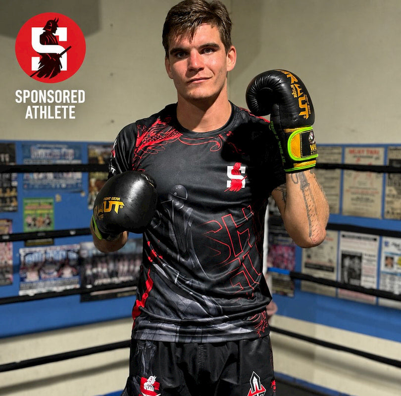A male athlete in a boxing ring wearing a black and red athletic jersey, black shorts, and black boxing gloves with yellow and green accents. The background shows a boxing gym with posters and a blue padded wall.