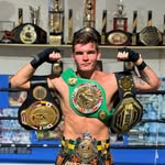 A male fighter in a boxing ring wearing a green and gold championship belt, holding up two gold and silver boxing championship belts, with a row of trophies on the wall behind him.