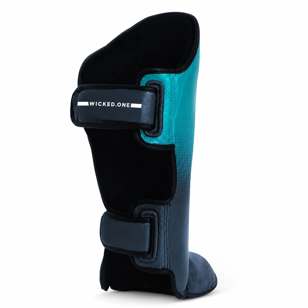 A single shin guard shown from the side on a clean white background. The product features a black body with a teal-to-dark-blue gradient panel and matching dark blue straps.