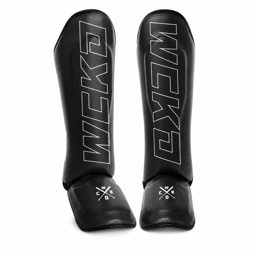 A pair of black shin guards with white outlined 'WCKD' branding on the shin area and a small WCKD logo on the foot cover, shown against a plain white background.
