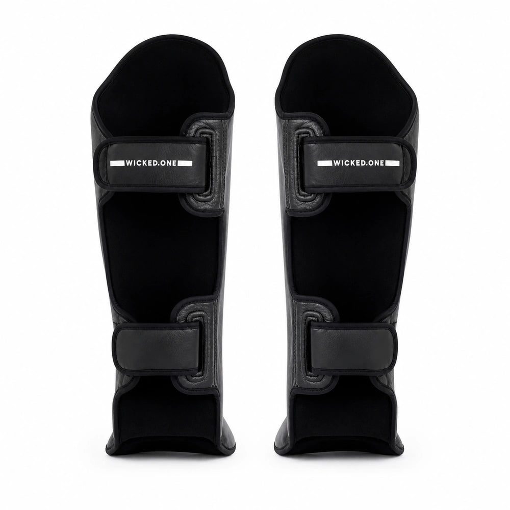 A pair of black shin guards with adjustable hook-and-loop straps, shown from the front on a solid white background. The brand name 'WICKED1'.