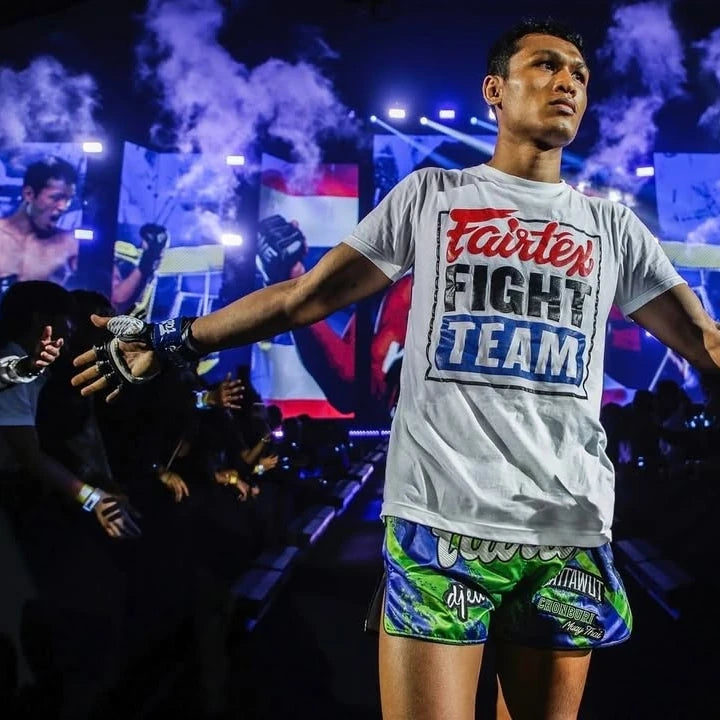 Person wearing a 'Fairtex Fight Team' t-shirt at a boxing event with smoke and lights in the background.