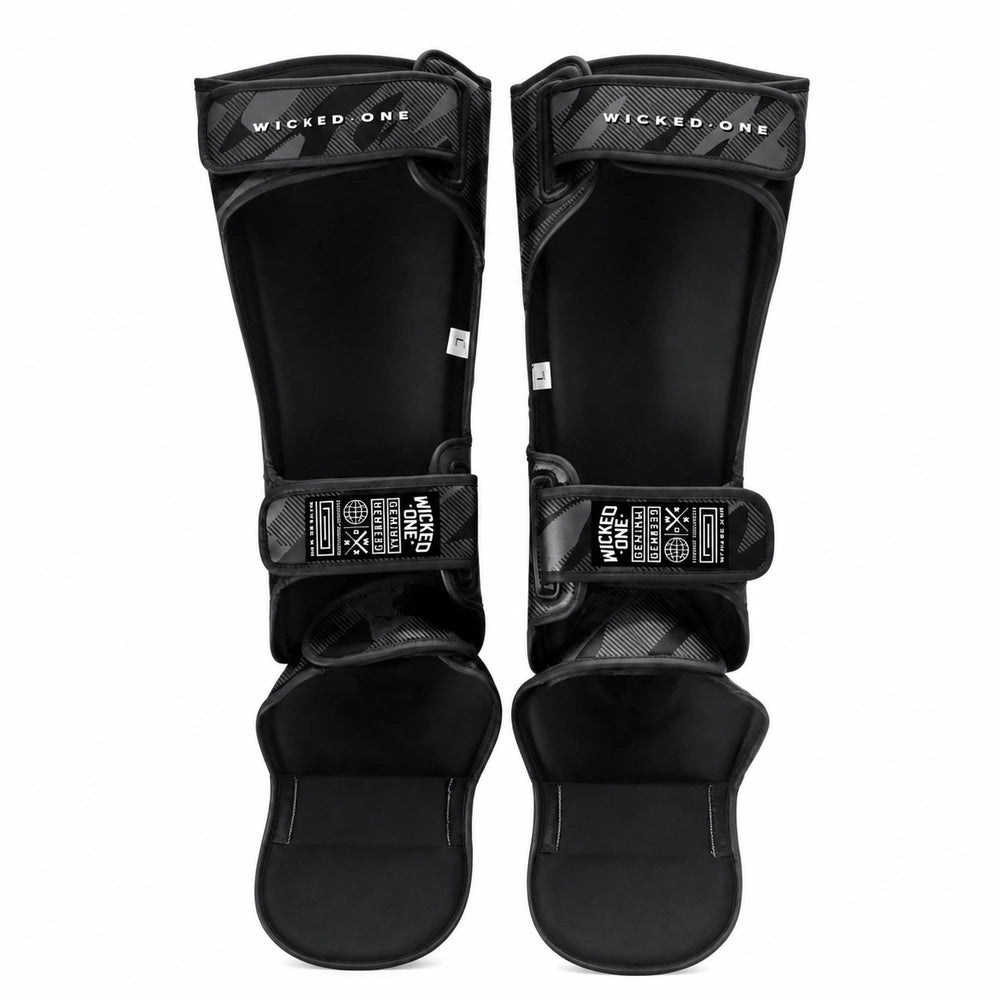 A pair of black shin guards with adjustable hook-and-loop straps and a carbon fiber pattern on the top straps, shown against a plain white background.