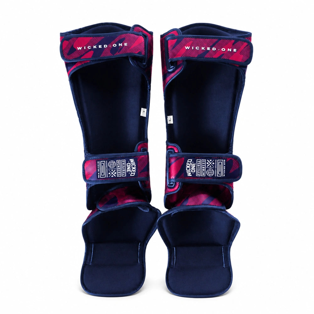 A pair of shin guards with navy blue velvet-like material and red/pink camouflage straps, featuring the brand name 'WICKED ONE' on the straps, shown against a plain white background.