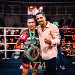 Two men in a boxing ring with one holding a championship belt, surrounded by boxing equipment.